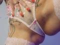 Close-up of ElixirShanna's lower torso. She's wearing sheer pink lace panties with floral embroidery, slightly pulled down to reveal her large, sagging breasts. Her hand with red-painted nails rests on the fabric. She has tattoos on her side, including a snake and a heart. The lace edges and her tattoos are in soft pink and black. The image has a slightly purple-tinted light.