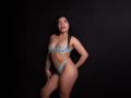 AdrianaPaul with long black hair, wearing light blue lace bra and thong, standing against dark background, showing tattoo on left arm, and smiling with red lipstick.