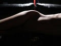 KamiLove's naked, pale-skinned, slender body is illuminated from the side, highlighting her smooth, curved ass and small, subtle, dark nipple on a black leather couch.