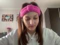 AdrianneEllinger, wearing a pink fuzzy headband, looking down with a slight smile, has brown hair, light skin, and is in a grey zip-up hoodie, showing small pimples on her face.