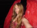 AkiGhil has long, wavy red hair, blue eyes, and is wearing a sparkly pink dress with red wings. Her tits are small, and she's taking a selfie.