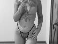 SugarrX in black-and-white mirror selfie, wearing a tied ribbed crop top and sheer lace panties, showing a belly button piercing and a tattoo on her right hip.