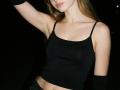 Diantrisy with small tits, wearing a black tank top, raised arm, black arm sleeves, and light skin, in dim lighting. Her long brown hair is down.