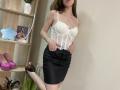 StellaNoir with white lace corset, black leather skirt, and pink heels, her tits pushed up, brown hair, hands on skirt, standing, a bookshelf with shoes and a photo.