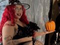 Estefy69 with red curly hair, glasses, black witch hat, black lace dress, black flower, holding orange pumpkin, tattoos on arm, smiling, dark lipstick, fishnet stockings, curvy, sexy.