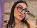 MaddyCarina with glossy lips, black glasses, and a nose ring, showing her tits and tattoos, wearing a gold necklace, with long dark hair, and a slight smile.