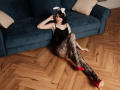ChrissieCandy with white bunny ears, black lace bodysuit, black lace stockings, and red high heels, sitting on wooden floor, arms behind head, blue sofa in background.