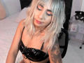 EmilyValk with blonde, wavy hair, wearing a black, shiny, leather bra, has a tattoo on her right arm, and is smiling with eyes closed. Boobs are visible.