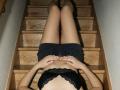 Destinis lying on wooden stairs, legs together, wearing black lace bra and black shorts, hands on stomach, black high heels, fair skin, visible lace bra edges, wooden stairs texture.