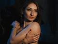 GwynBartell with red nails, crossed arms, and a serious expression, wearing gold earrings and a bracelet, in dim blue lighting. Her tits are partially covered, looking sexy.