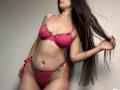Tisia with long brown hair, wearing pink lace bra and matching panties, holding hair to side, showing her tits and pussy, standing against a plain wall.