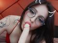 PaisleyMour with glasses, black hair, red top, tattoos, fellating a dildo. Dildo in her mouth, eyes looking up, red headboard in background. Her tits are visible.