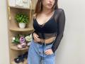 Rioria with brown hair, black sheer top, showing her tits, blue jeans, black belt, and gold earrings, standing in front of a wooden shelf with flowers and a plant.