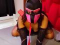 LilithAndAsmodeo in black mask with pink stripes, red leash, curly hair, black lace gloves, and dress, kneeling on bed, showing off her big ass and boobs.