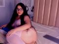 KonyBella with long black hair, glasses, and pink lace lingerie, laying on a bed, showing her big ass, wearing white thigh-highs, remote control on the bed.
