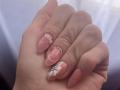 EliseMonet's hand with pink, almond-shaped nails, featuring white floral designs, small pink and gold accents, and a gold ring on the ring finger, clenched in a fist.