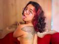 KanelaMiller with red and black curly hair, glasses, and tongue out, topless, showing a large tattoo on her back, red couch in the background.