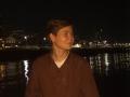 MarybelleDoerr with short brown hair, wearing a brown button-up shirt and white tee, gazing to the side at night, reflection in water, city lights in background.