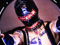 AmySpider with shiny black latex mask, red lace-up, tattoos on chest and arms, black tank top, and black gloves, holding up camera, in a high-contrast, circular-shot image.