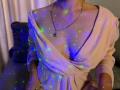 Anshante with glowing green and blue dots on her pale skin, wearing a pink, deep V-neck dress, silver necklace, and no visible tits or ass. She's in a dimly lit room.