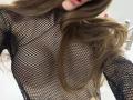 SensualMila's face is partially visible with long brown hair. She's wearing a black fishnet top, revealing her small, perky tits through the net. Her lips are slightly open.