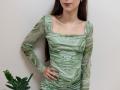 AmeliaMaiv in a green marble-patterned dress, standing with hands on hips, long brown hair, light skin, small tits, slim build, slight smile, small plant on the left.