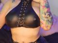 Kkole's big tits in a shiny black leather halter top with lace-up front, showing her tattooed arm with colorful cat design and blue eyes. Her hand touches her neck.