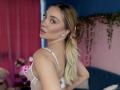 JessicaClarks with blonde ponytail, looking back, wearing white sheer lingerie, showing small tits, pink lips, blue eyes, and subtle makeup, in a colorful bedroom.