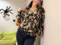 Tilliana, com os seios levemente visíveis sob uma blusa floral marrom, vestindo jeans escuros, encostada em uma parede, cabelos castanhos, óculos, mãos na cintura, em um quarto com tema de Halloween..