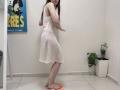 JoannaSia in a white, see-through dress with her ass and pussy slightly visible, standing with arms out, orange ring on floor, potted plant, and wall art in background.