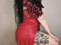 Niferttiti in a red sheer lingerie dress with black roses, lace, and floral tattoos on her thigh, black curly hair, and black lace mask, posing with her ass up.