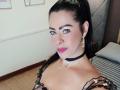 SofiMontes with black hair in a ponytail, wearing pink lipstick, black choker, gold earrings, and sequined top, showing her tits and a necklace, in a bedroom.