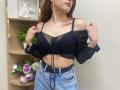 Rioria with small tits, wearing a black sheer off-shoulder top and blue denim high-waisted jeans, holding top straps, brown hair, gold hoop earrings, standing in a room.