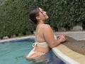 LorensDouce with her tits and ass in a white bikini, standing in a pool, eyes closed, leaning on the edge, brown hair in a bun, hoop earrings, water around her.