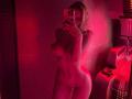 Nude MillieHartwell, red-lit, stands, covering her tits with one hand, holding a phone to her face, her ass and pussy visible, in a mirror selfie.