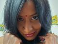 Nahirr with dark, wavy hair, a red bindi on her forehead, and a nose ring, smiling with red lipstick, hands near her face. Her eyes are dark and expressive.