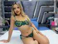 CloeRouss with blonde hair, green lace bra and panty set, sitting on a bed, showing her tits and ass, red lipstick, tattoo on right arm, confident smile.
