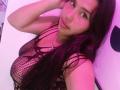 GemmaBellini with long black hair, wearing a black fishnet top, showing her large tits, pinkish-purple light, looking at the camera with a slight smile, hand on her head.