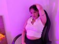 BelaDeimons with medium tits, wearing a white ribbed sleeveless top, black chair, right arm raised, purple-lit room, black skirt, tattoos on arm, looking at camera.