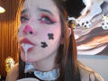 AryButty with cat ears, pink nose, black makeup dots, white fluid dripping from mouth, pink eyelids, brown hair, wooden wall, soccer ball decor, and a lamp in background.