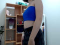 Steisii with light skin, blue strapless top, black jeans, visible small ass, short brown hair, standing, mirror reflection, wooden shelf with pink toy and potted plant in background.
