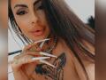 DenyChantal with long brown hair, thick eyebrows, long white nails, tattoos on chest, necklace, and full pink lips, showing off her tits and hand tattoos.