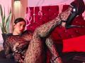 DiannaRosse in black lace snakeprint bodysuit, showing her crotch and pussy through the fabric, legs up in black stilettos, on a red tufted bed, pink wall in background.