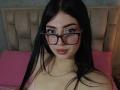IsabellaaRossi with black glasses, pink lipstick, and long black hair, wearing a pink top, sitting on a pink bed, showing her tits, with a silver necklace.