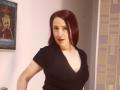 SweettEve with red hair, black V-neck shirt, small tits, pale skin, and red lipstick, standing in a room, wall with patterned wallpaper on the right.