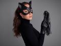 AmmaLoyXX with cat mask, black turtleneck, and leather gloves holding a flogger, her brown wavy hair in a ponytail, looking back with a confident expression.