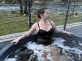KarKutie with wet black dress, tattoos on arms, small tits, and brown braided hair, sits in a bubbling black hot tub, looking to the side.
