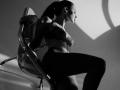MilagrosEdmisten with dark hair in a ponytail, wearing a lace bra and high-waisted pants, sitting on a clear chair, shadows on the wall, looking to the side.
