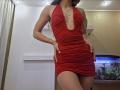 MilaYanis wearing a red ruched halter dress with a deep cleavage, hands on hips, showing her slim figure and small ass, in a brightly lit room.