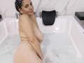MadameScarlett is naked in a bathtub with bubbles, showing her big tits and ass, smiling with wet skin, dark hair in a headscarf, and black seat behind her.